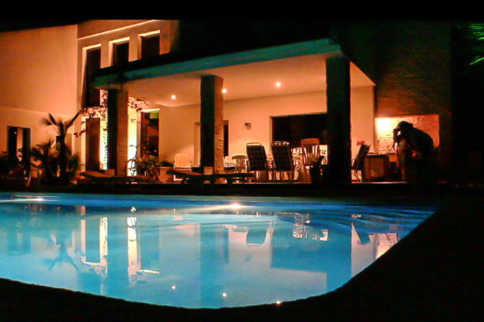 Terrasse couverte éclairée et piscine privée de nuit, avec des piliers en pierre et un espace repas extérieur sous un éclairage chaleureux.
