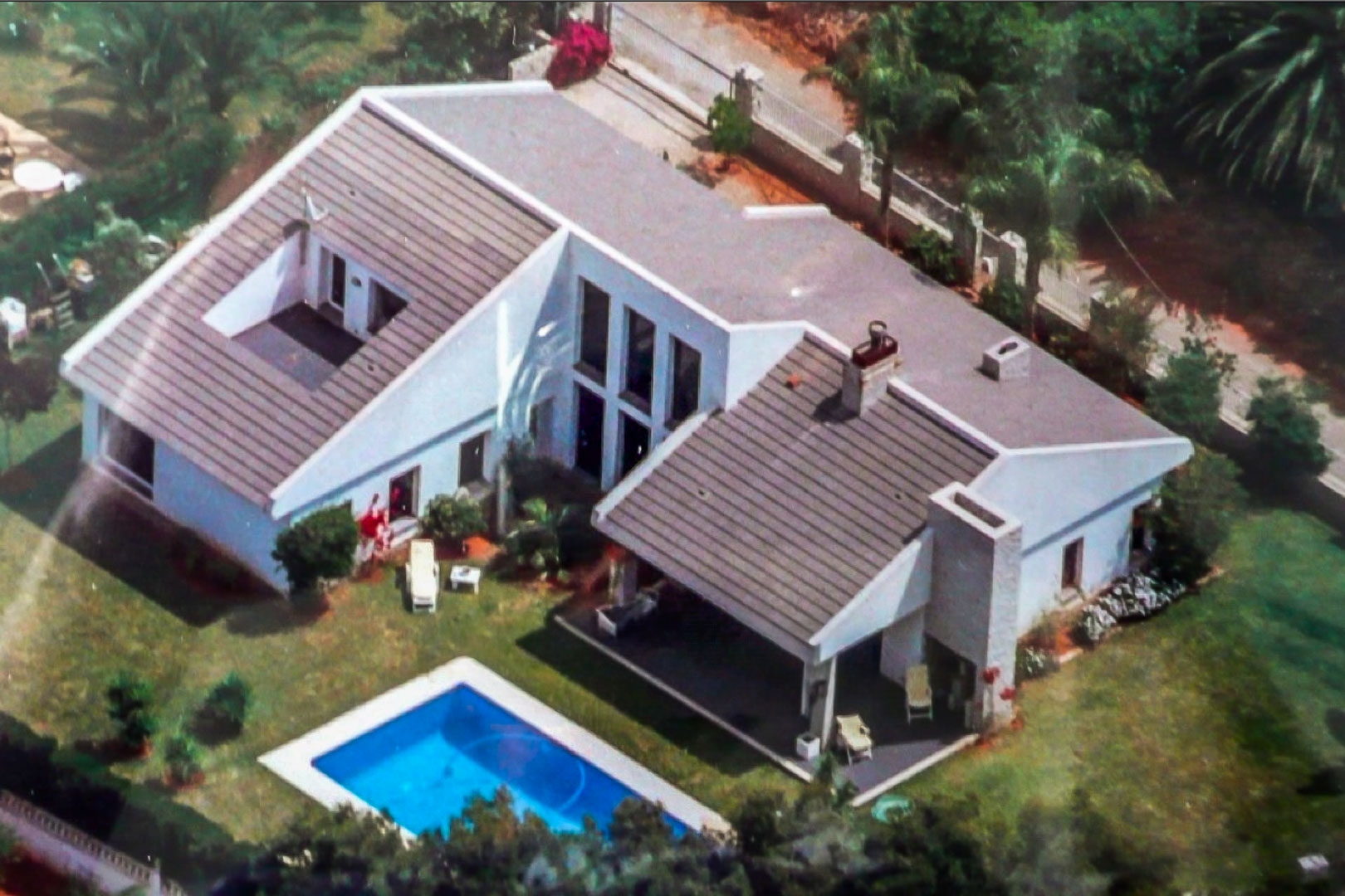 Vue aérienne de la villa blanche avec toit en tuiles, piscine rectangulaire et jardin paysager comprenant une terrasse couverte et une clôture de protection.