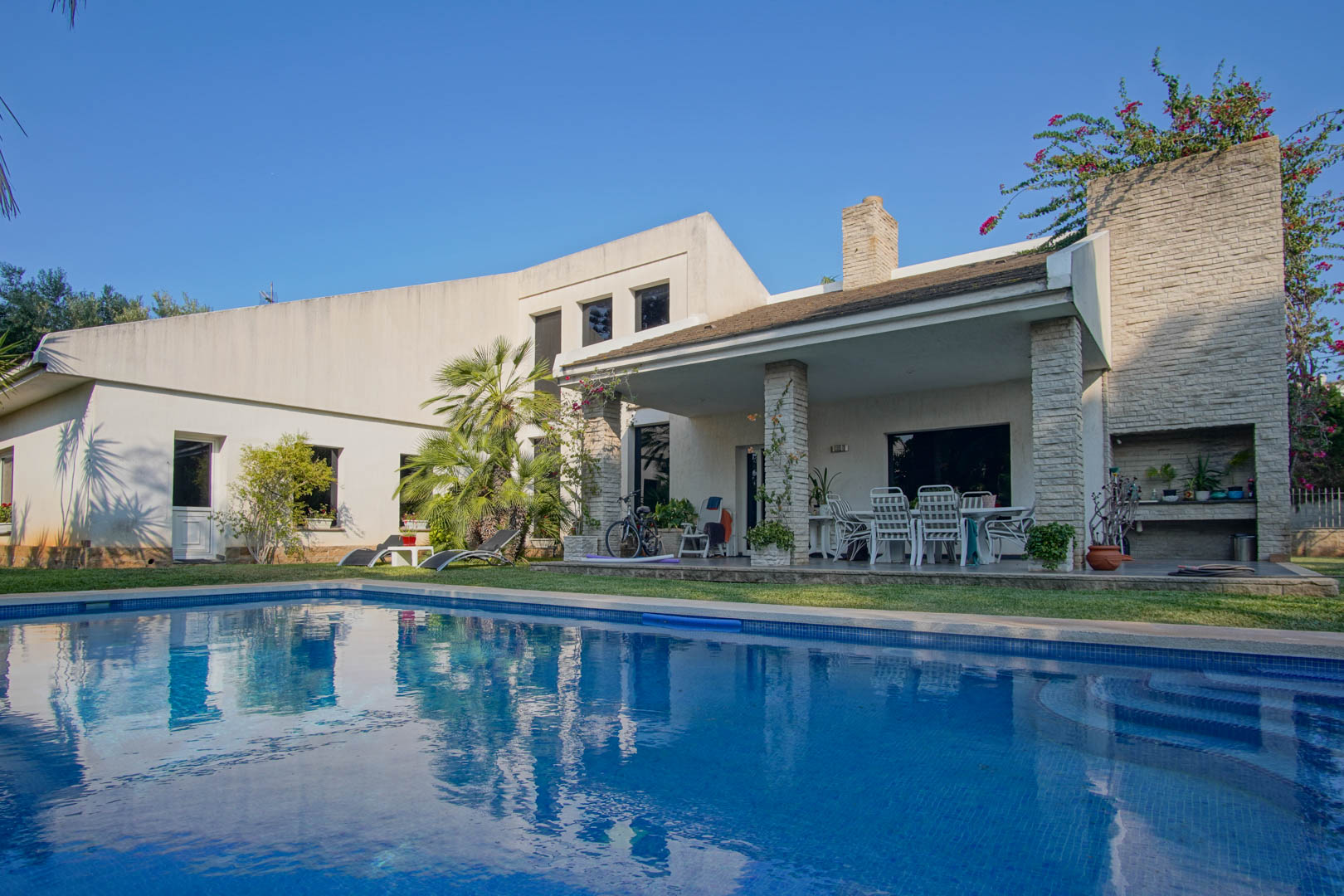 Vue extérieure de la piscine privée et de la terrasse couverte avec piliers en pierre, comprenant un coin repas et un espace barbecue intégré.