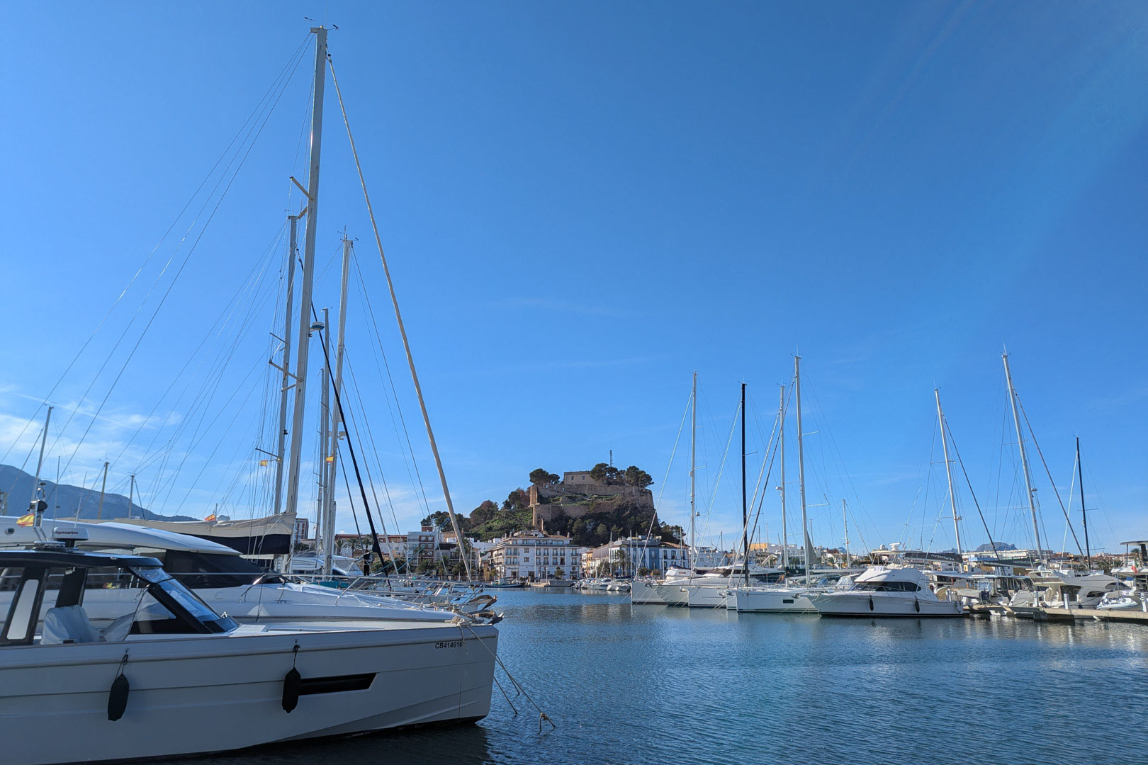 Vue panoramique du port de plaisance de Dénia avec ses yachts et le château historique dominant la ville sous un ciel bleu azur.