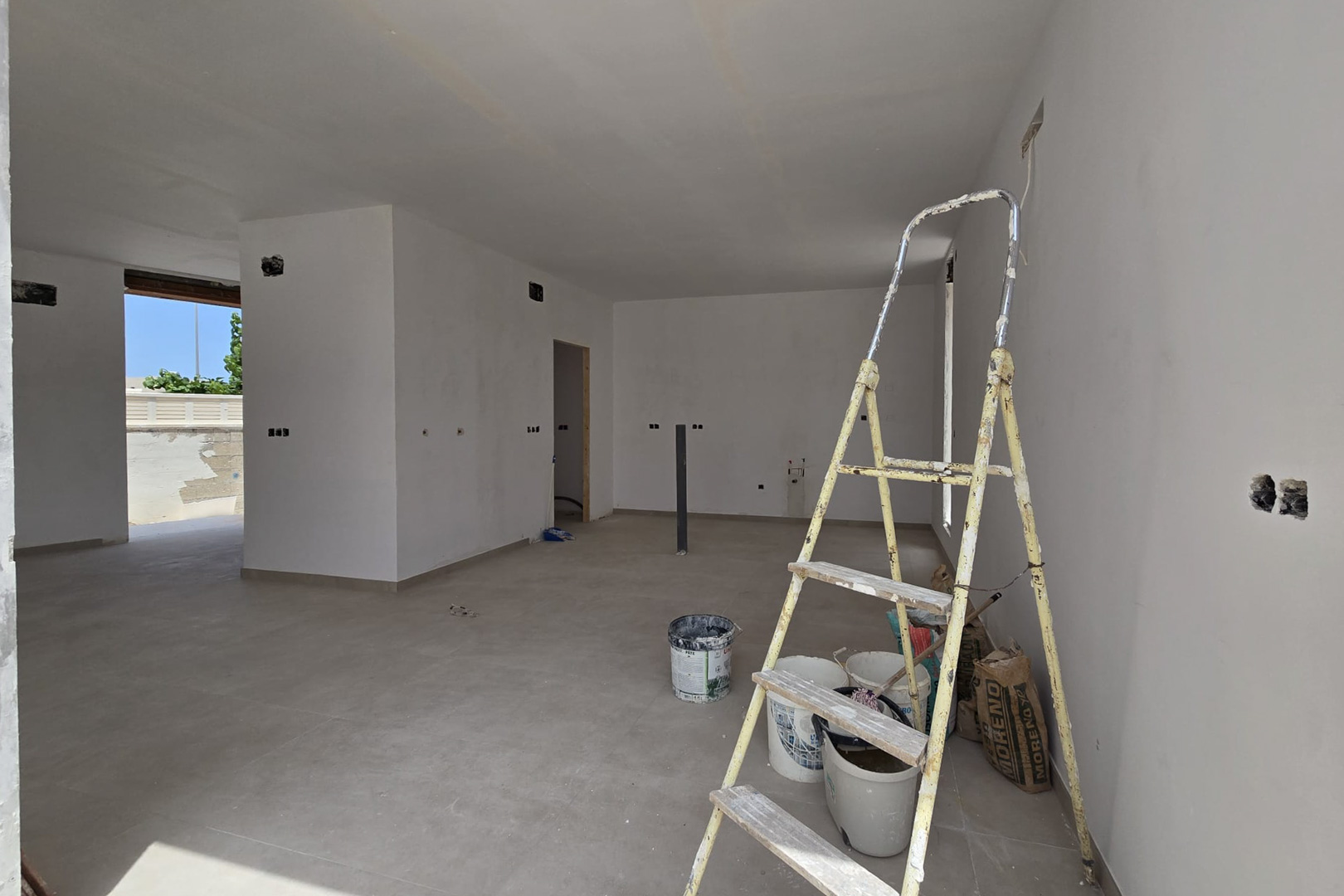 Intérieur à aire ouverte en phase de finition avec sol en céramique grand format et murs blancs. On aperçoit les pré-installations électriques et l'accès extérieur.