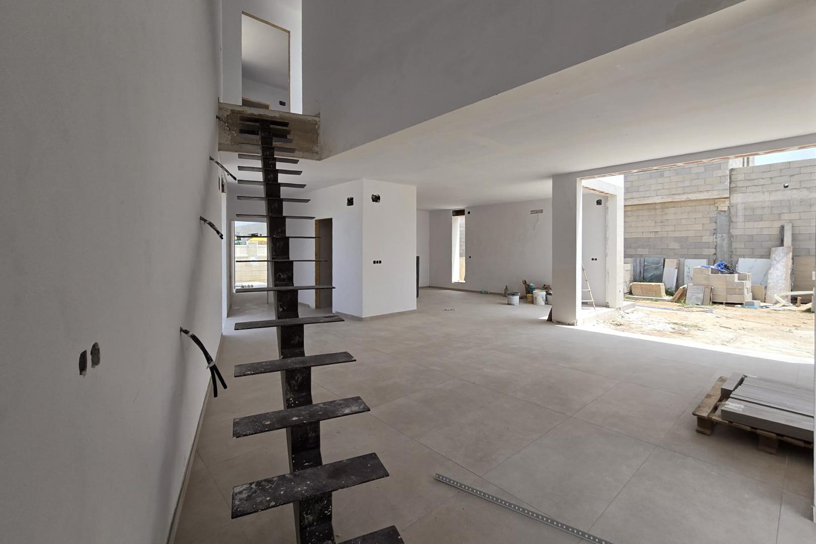 Intérieur en construction avec escalier métallique suspendu, carrelage grand format et mezzanine offrant une belle hauteur sous plafond et de la lumière.