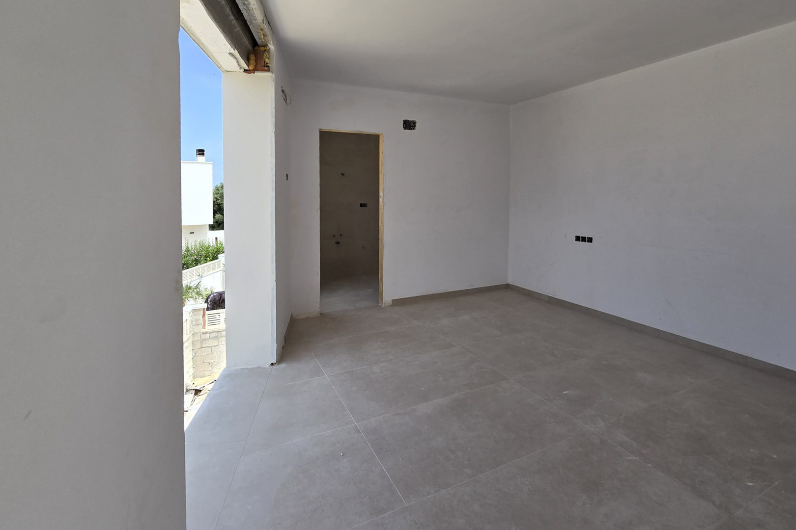 Pièce intérieure en construction avec carrelage gris grand format, murs blancs et accès extérieur. On aperçoit l'entrée d'une salle de bain attenante et les installations électriques.