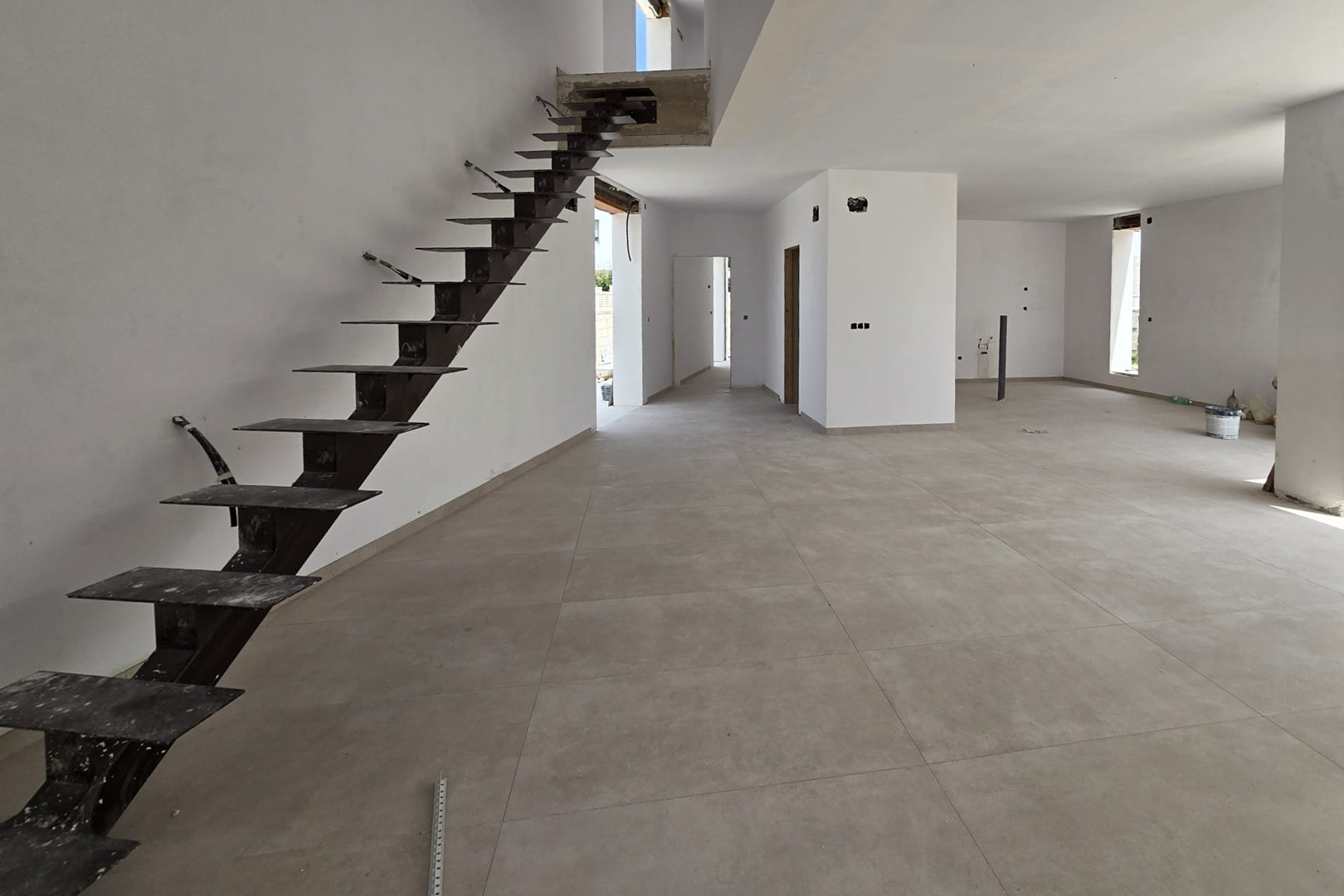 Rez-de-chaussée à aire ouverte en construction avec carrelage en grès cérame gris et un escalier suspendu à structure métallique noire.