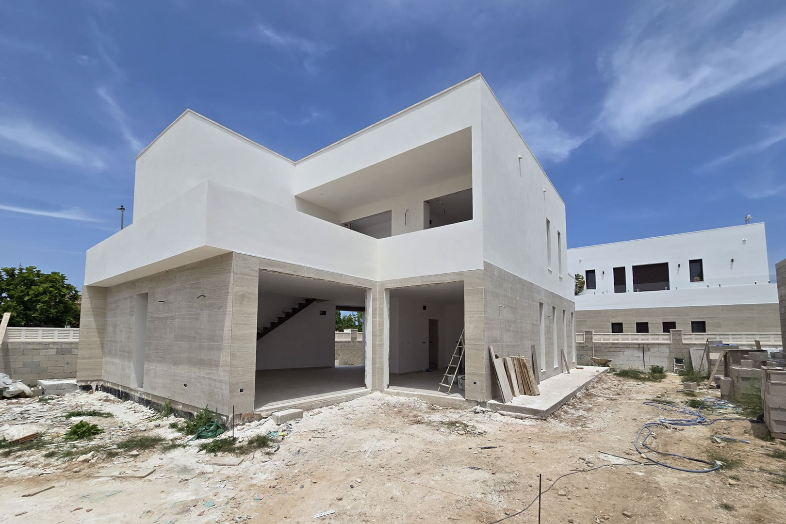 Vue extérieure de la villa en construction avec façade en pierre au rez-de-chaussée, volumes blancs à l'étage et larges ouvertures pour baies vitrées.