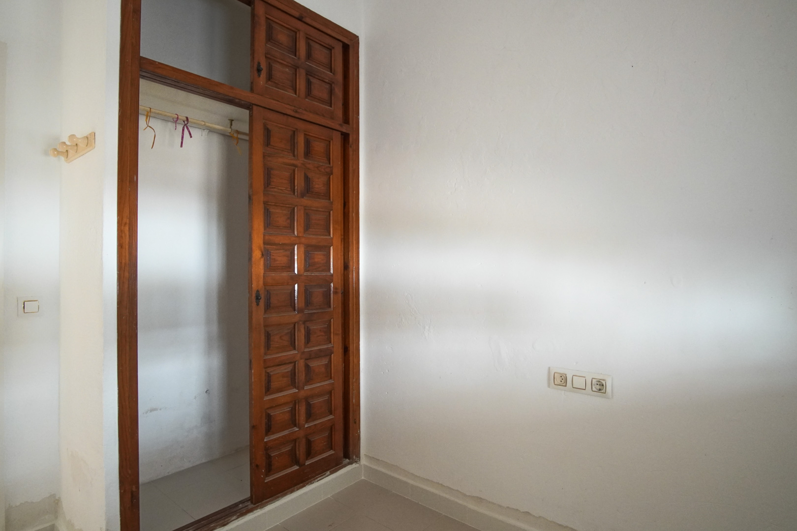 Chambre avec placard intégré en bois foncé et portes coulissantes à panneaux. Les murs blancs texturés et le sol en grès complètent cet espace fonctionnel.