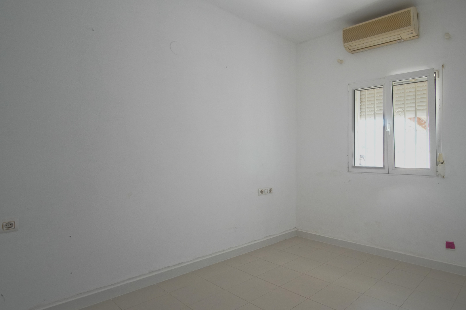 Chambre lumineuse avec murs blancs, sol en grès et climatisation murale. Elle dispose d'une fenêtre en PVC avec volet roulant et de plusieurs prises électriques.