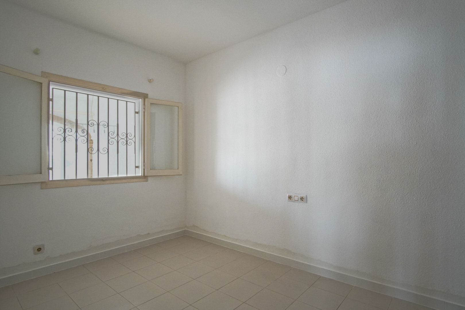 Chambre lumineuse avec murs blancs texturés, sol en grès cérame clair et fenêtre extérieure équipée de grilles de sécurité décoratives.