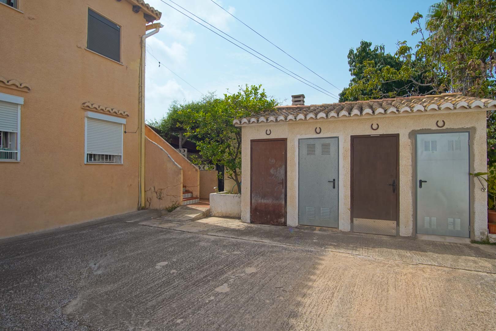 Espace extérieur pavé comprenant une annexe fonctionnelle avec quatre débarras individuels, surmontés de fers à cheval décoratifs sous un toit en tuiles.