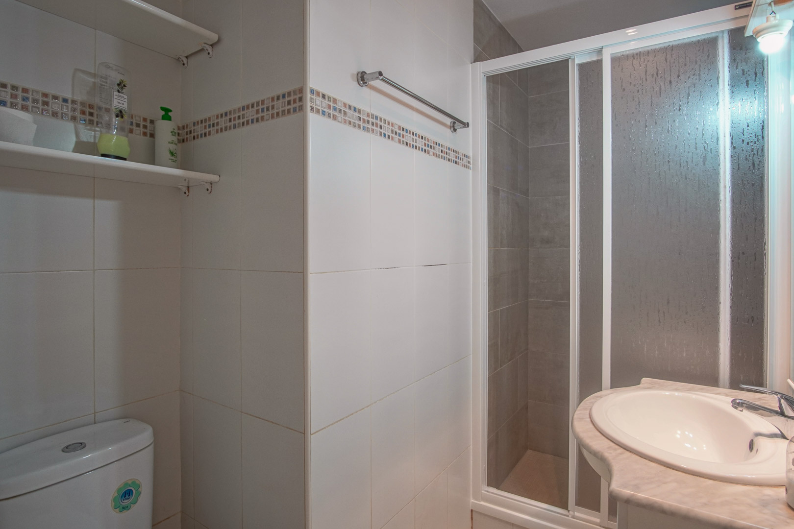 Salle de bain aux murs carrelés blancs avec frise décorative, comprenant une douche à paroi coulissante et un lavabo intégré dans un plan de travail en marbre.