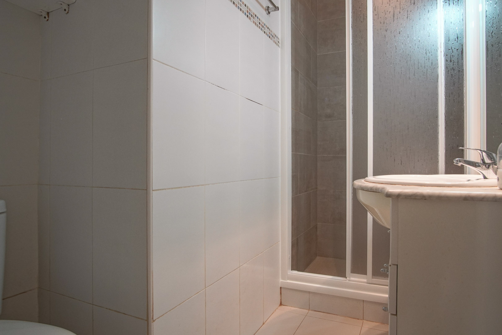 Salle de bain fonctionnelle avec murs carrelés blancs, cabine de douche à paroi givrée et lavabo encastré sur un plan en marbre.