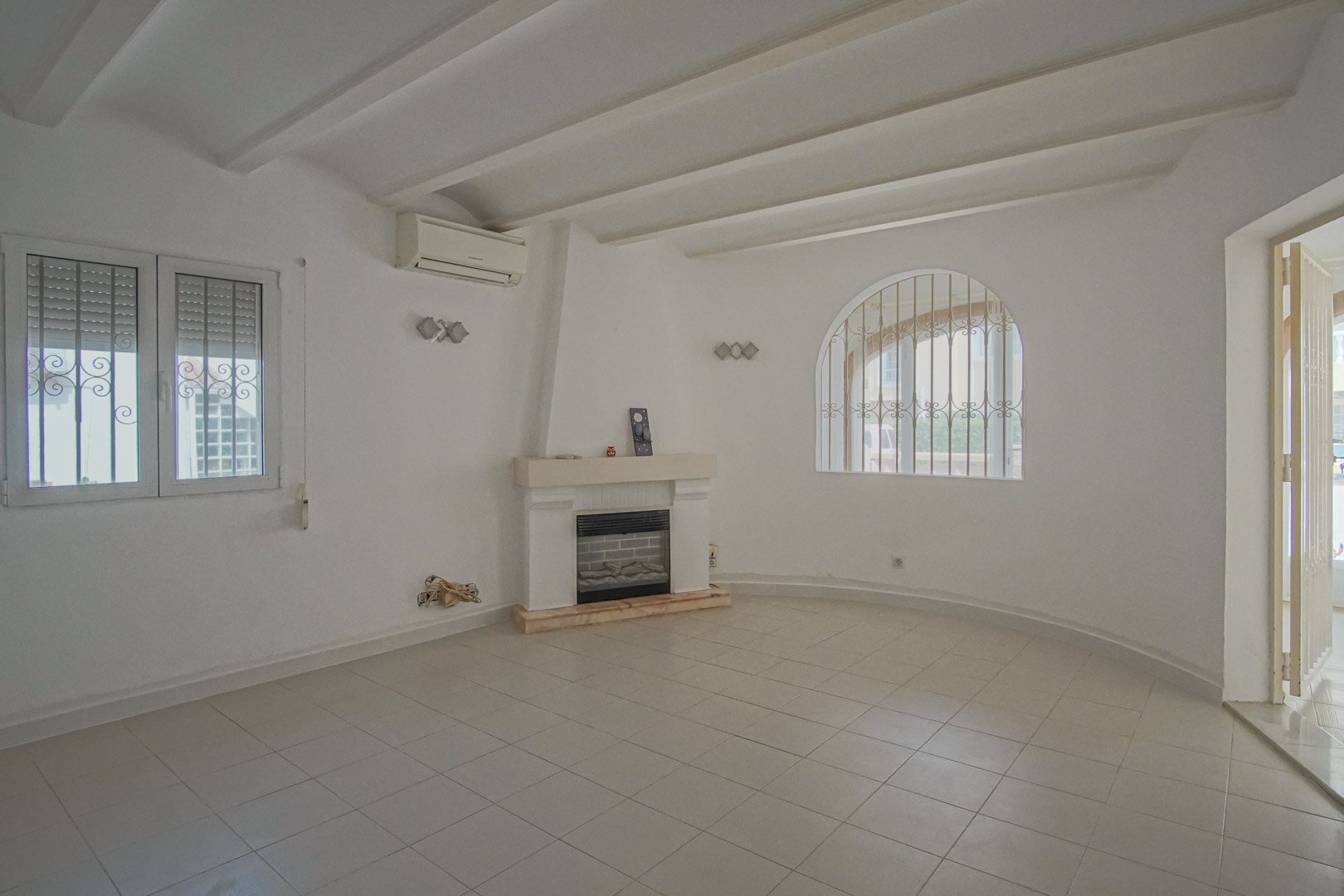Salon avec cheminée centrale, plafond à poutres apparentes blanches et fenêtre en arche avec grilles décoratives. Équipé de la climatisation et d'un sol en grès.
