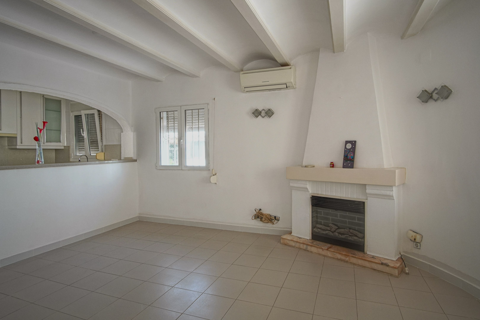 Salon avec cheminée, plafond à poutres blanches et climatisation. Une ouverture en arche relie cet espace de vie à la cuisine semi-ouverte.