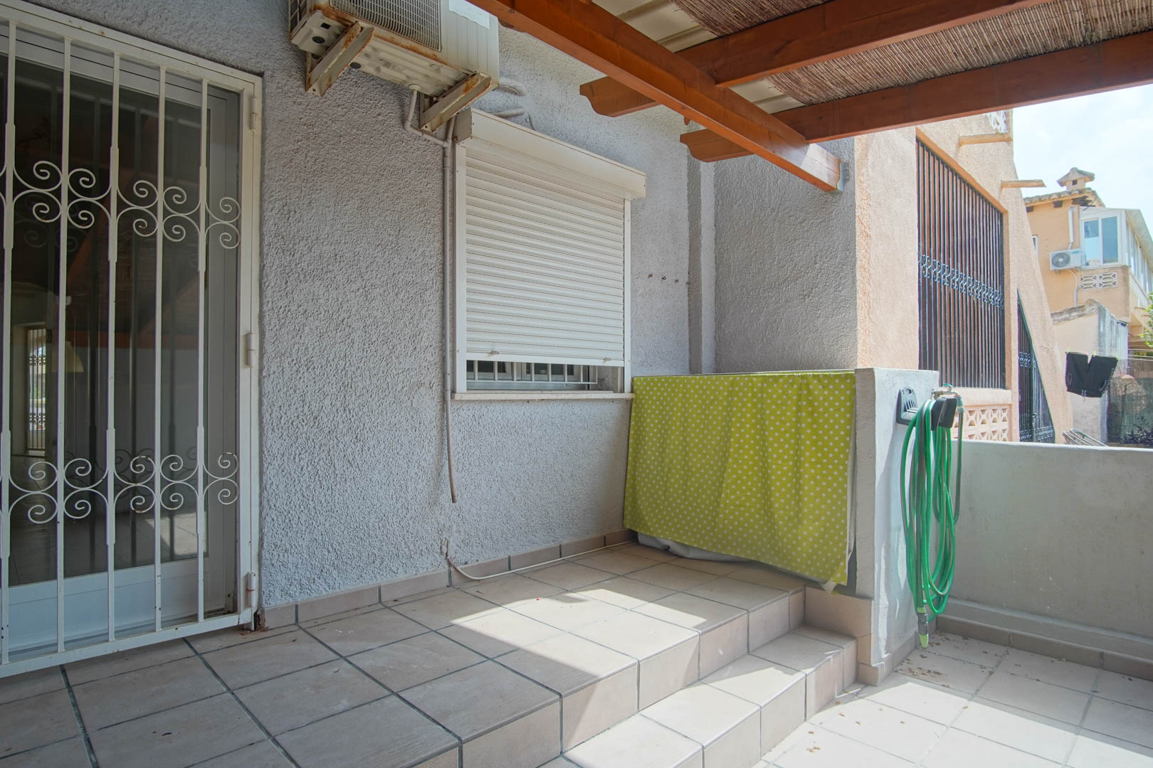 Terrasse extérieure avec sol en grès, pergola en bois avec canisse et porte sécurisée par une grille. Comprend une unité de climatisation et un espace utilitaire.
