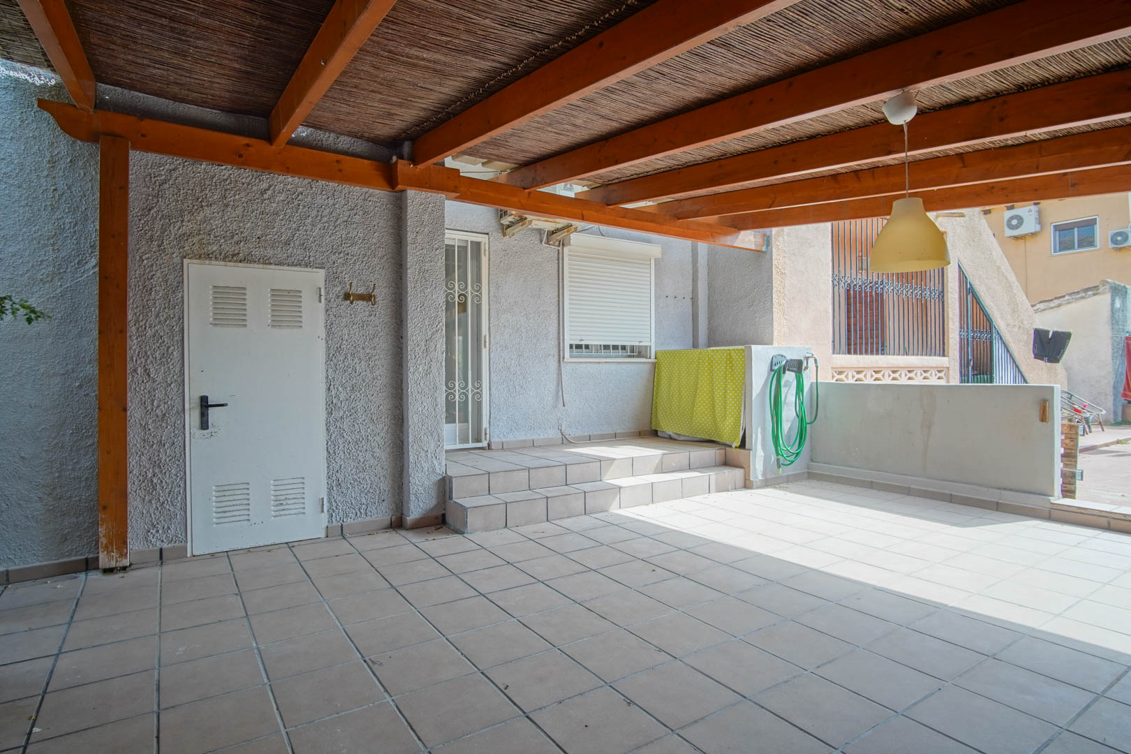 Terrasse extérieure couverte par une pergola en bois et canisse, dotée d'un sol en carrelage et d'un accès direct à l'habitation par des marches et une porte sécurisée.