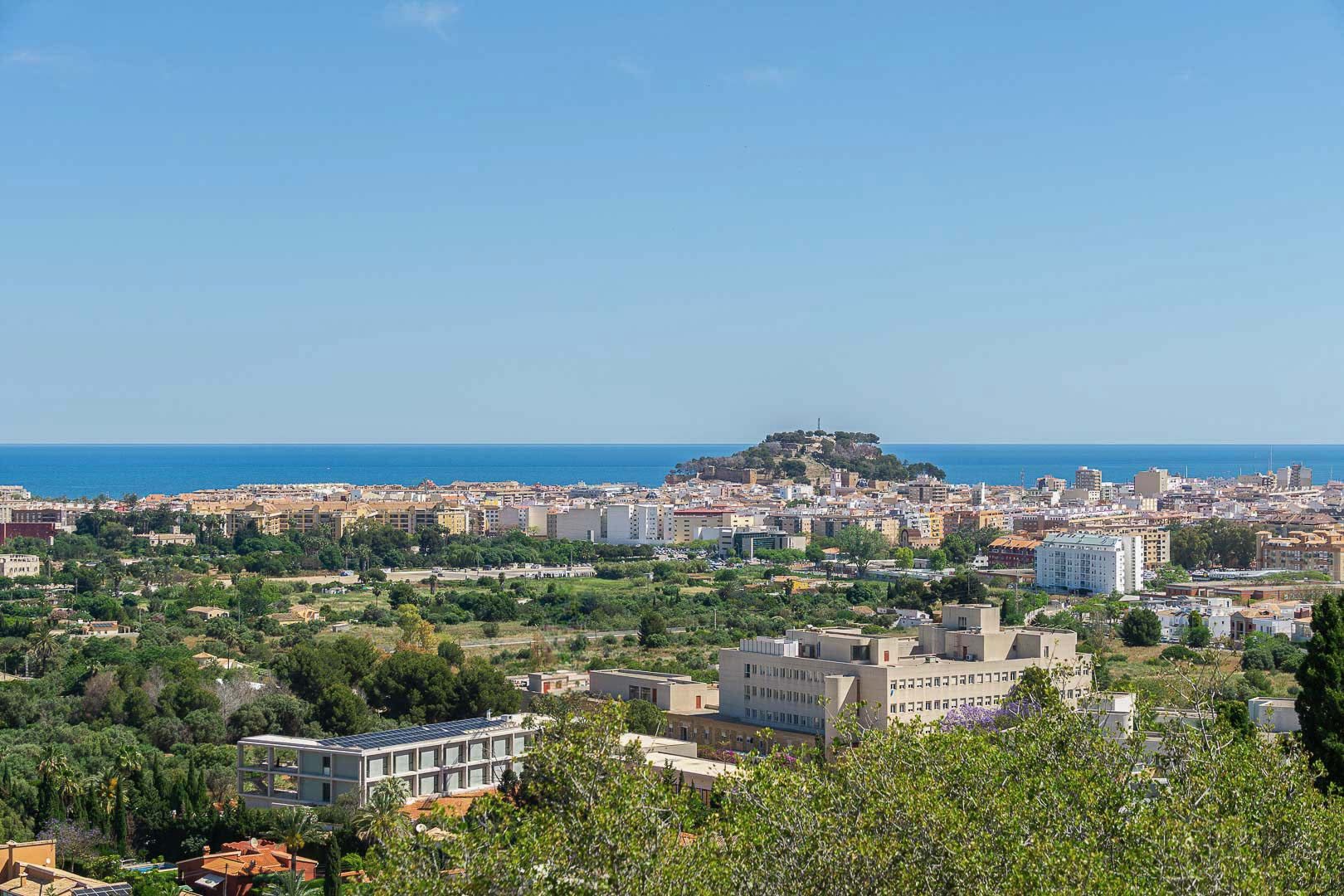 Vue panoramique de Dénia avec son château historique sur la colline, la mer Méditerranée à l'horizon et le paysage urbain entouré de verdure.