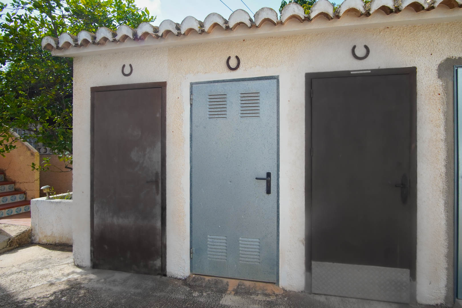 Zone de rangement extérieure comprenant trois portes métalliques sous un toit en tuiles traditionnelles, ornée de fers à cheval décoratifs.