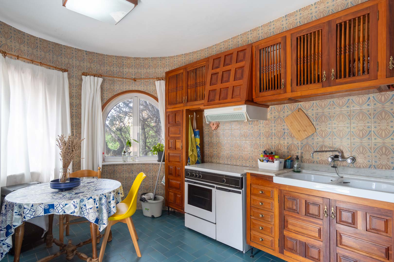 Cuisine rustique avec meubles en bois sculpté, carreaux muraux à motifs et coin repas devant une fenêtre en arc offrant une vue sur la végétation.