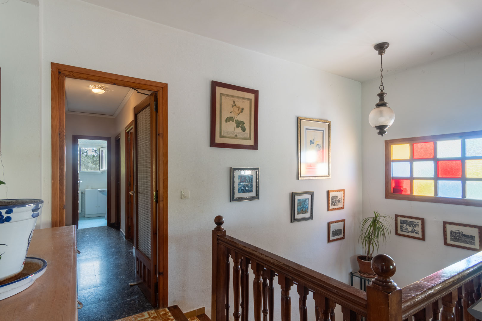 Palier de l'étage supérieur avec balustrade en bois tourné, vitrail coloré et couloir menant aux chambres avec encadrements de portes en bois naturel.