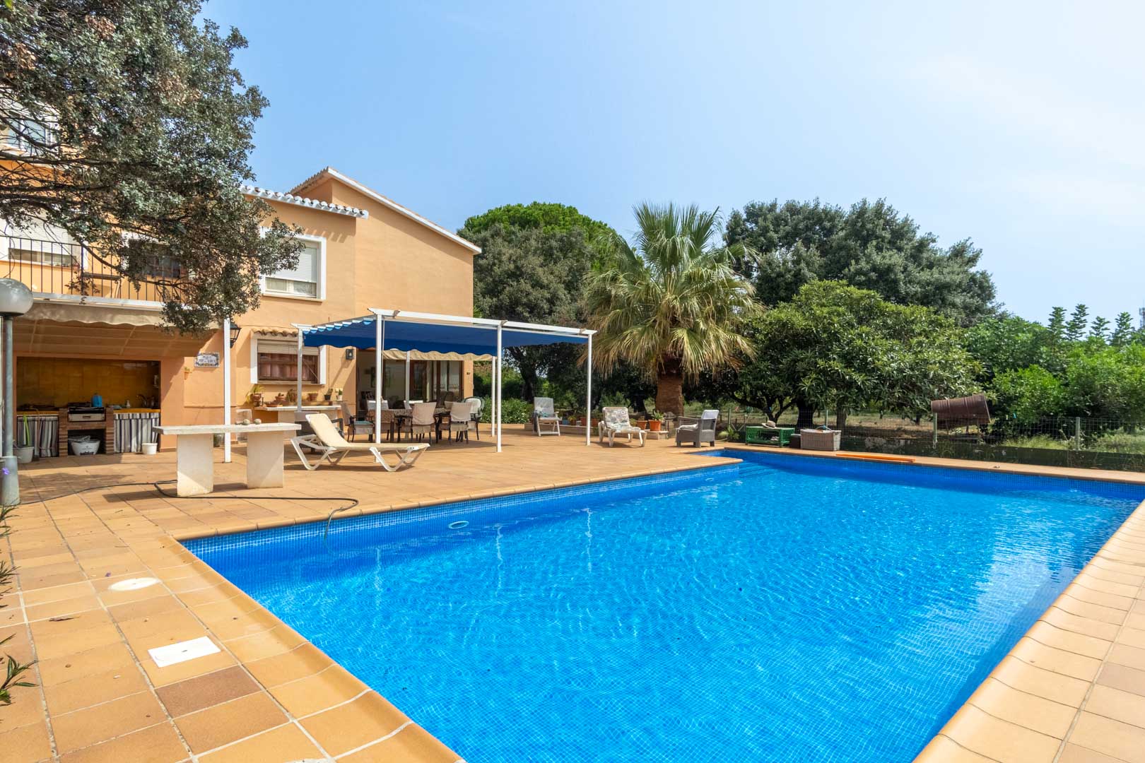 Piscine rectangulaire en mosaïque bleue entourée d'une terrasse en terre cuite, avec cuisine d'été et espace dînatoire ombragé par un store bleu.