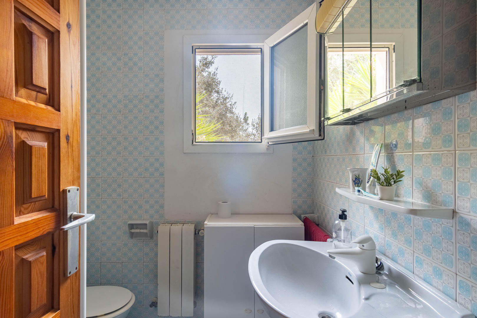 Salle de bain avec carrelage mural à motifs bleus, lavabo blanc et fenêtre oscillo-battante offrant une luminosité naturelle et une vue sur l'extérieur.