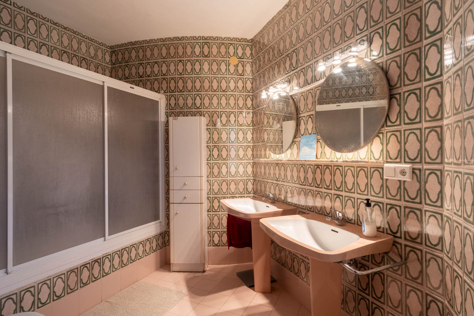 Salle de bains au style rétro avec double vasque, miroirs circulaires éclairés et carrelage à motifs verts. Comprend une douche à parois givrées et une colonne de rangement.