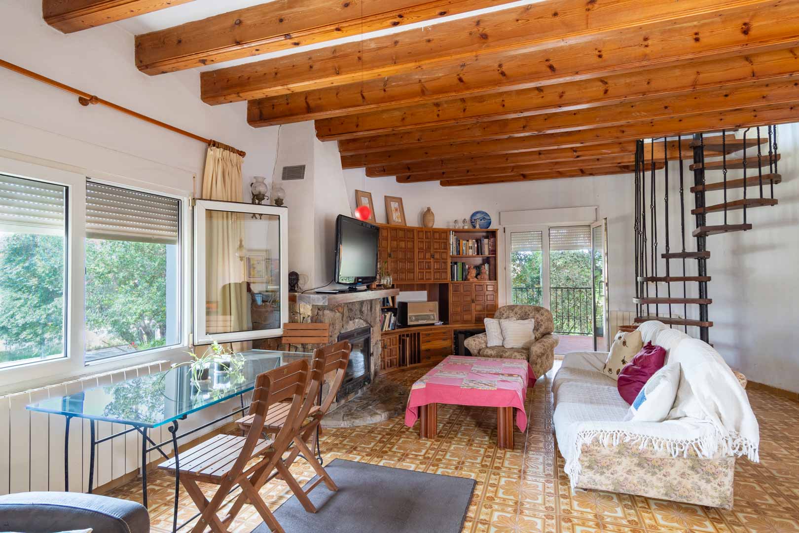 Salon avec poutres apparentes en bois, cheminée en pierre et un escalier en colimaçon en fer forgé menant à l'étage supérieur.
