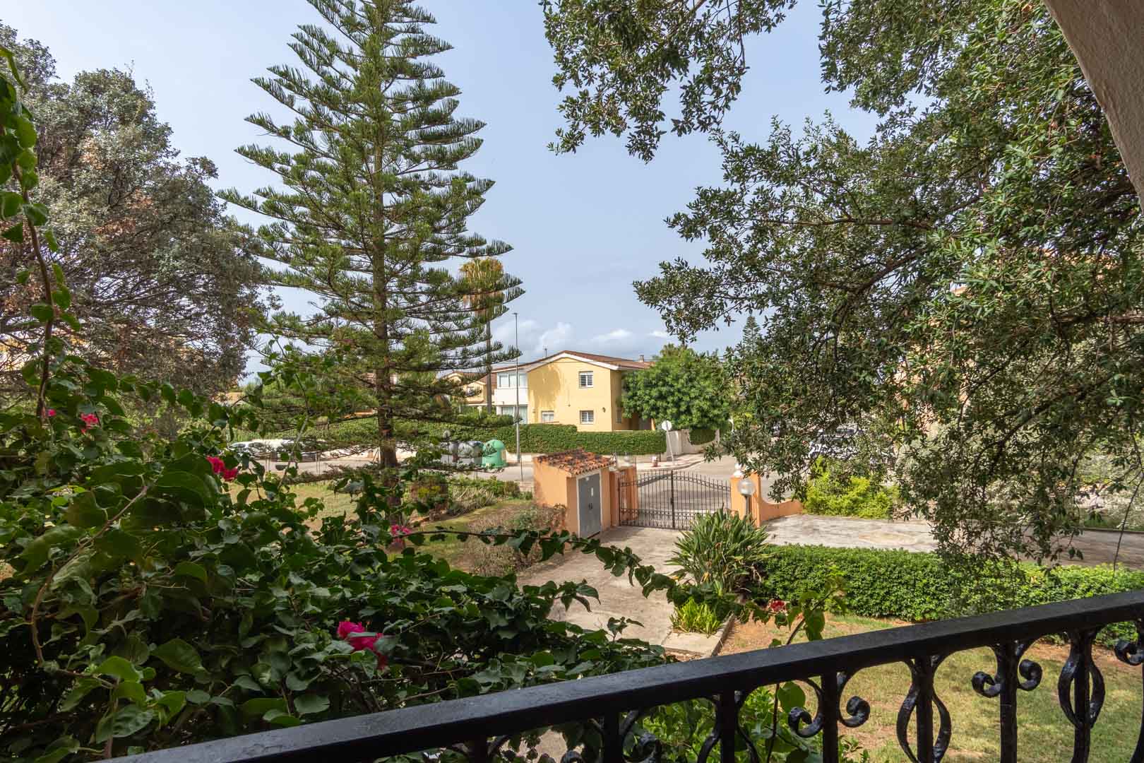 Vue depuis la terrasse supérieure avec garde-corps en fer forgé, donnant sur le jardin méditerranéen arboré et le portail d'entrée de la propriété.