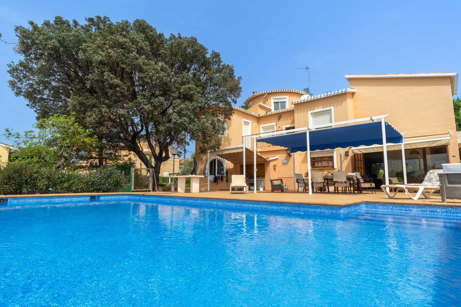 Vue extérieure de la piscine privée et de sa terrasse, mettant en valeur la façade méditerranéenne de la villa et son espace d'été ombragé par un store bleu.