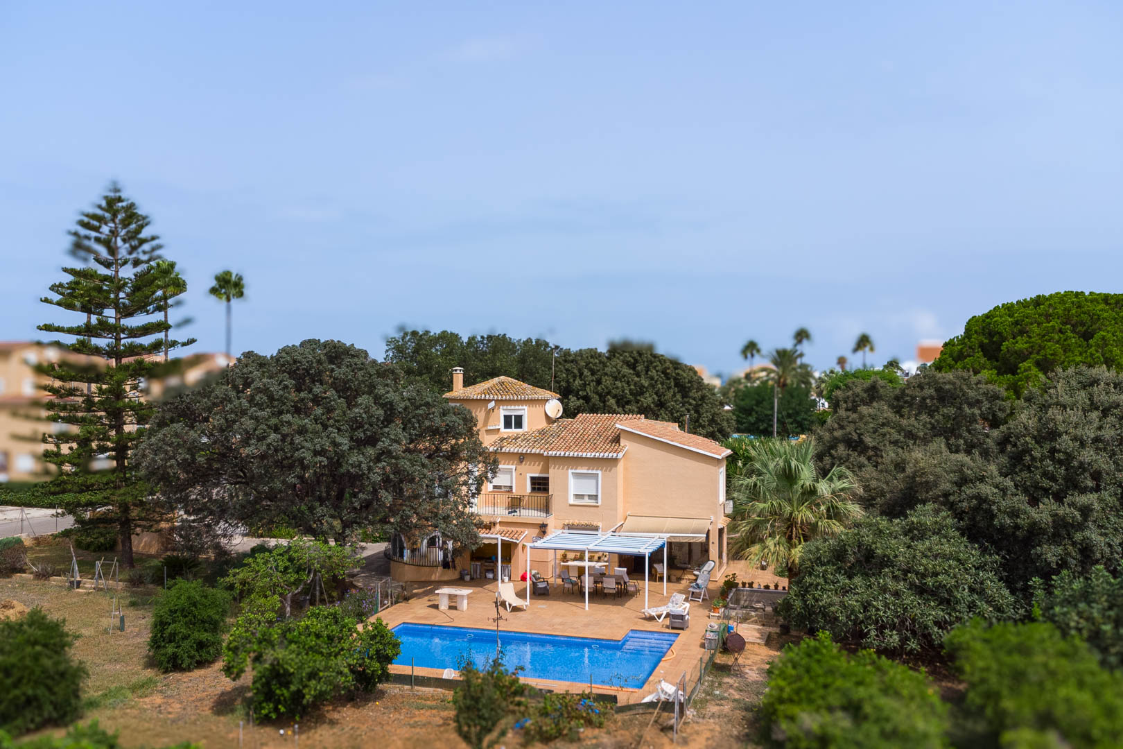 Vue extérieure de la villa avec piscine rectangulaire, terrasse sous pergola blanche et coin repas, le tout entouré d'une végétation méditerranéenne et d'arbres matures.