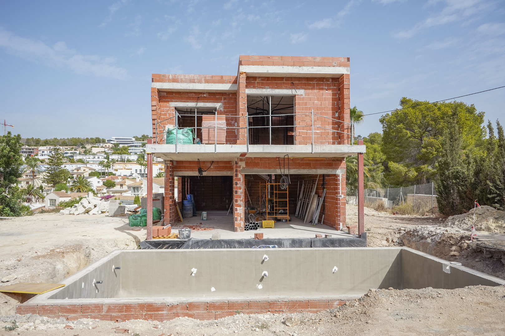 Phase de construction actuelle de la villa présentant la structure en briques sur deux niveaux et le bassin de la piscine en béton au premier plan.