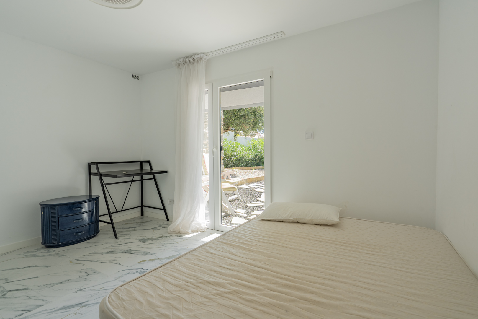 Chambre avec sol en marbre et accès direct à la terrasse du jardin. Équipée d'un bureau noir et d'une commode bleue sous une lumière naturelle.