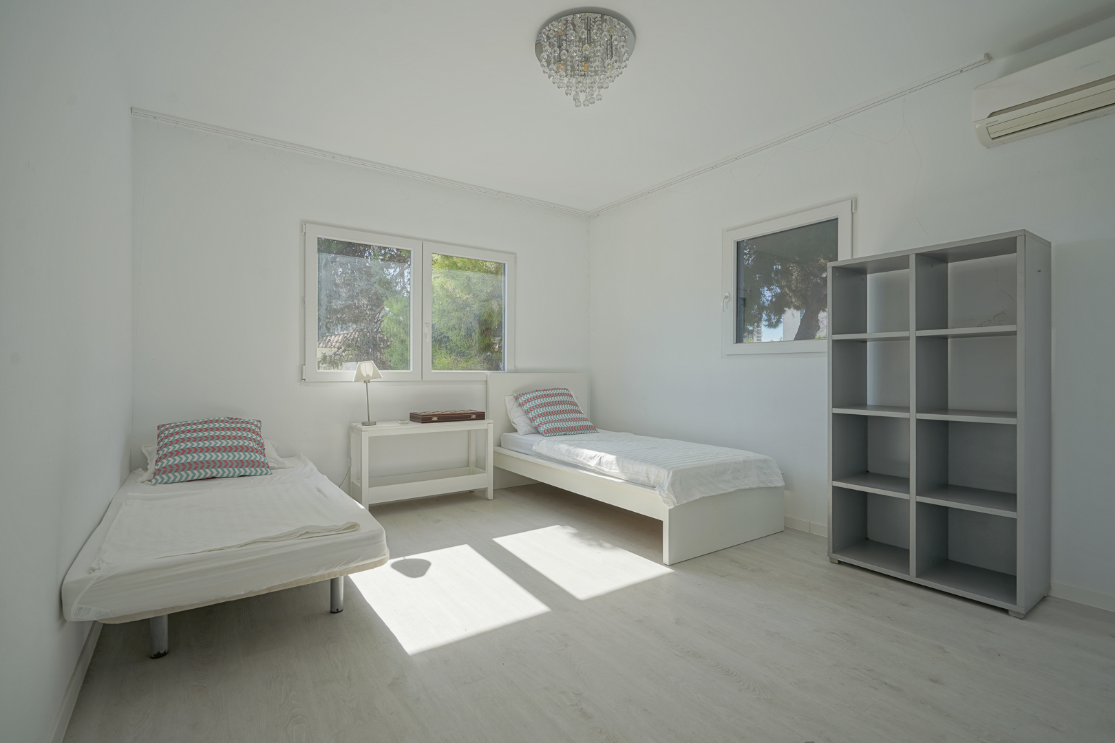 Chambre lumineuse avec deux lits simples, parquet clair et étagère grise. Équipée de la climatisation, d'un lustre en cristal et de fenêtres offrant une vue sur la végétation.