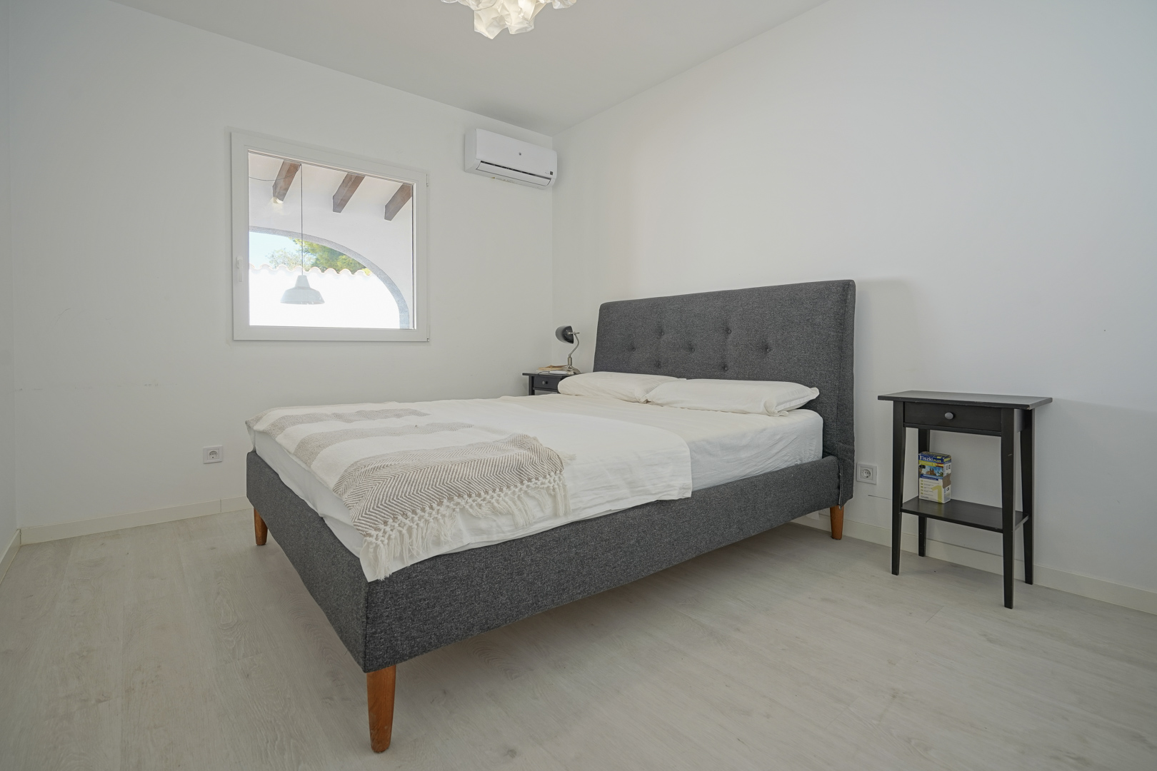 Chambre lumineuse avec lit double rembourré gris, climatisation réversible et sol stratifié clair. La fenêtre donne sur une terrasse couverte aux poutres apparentes.