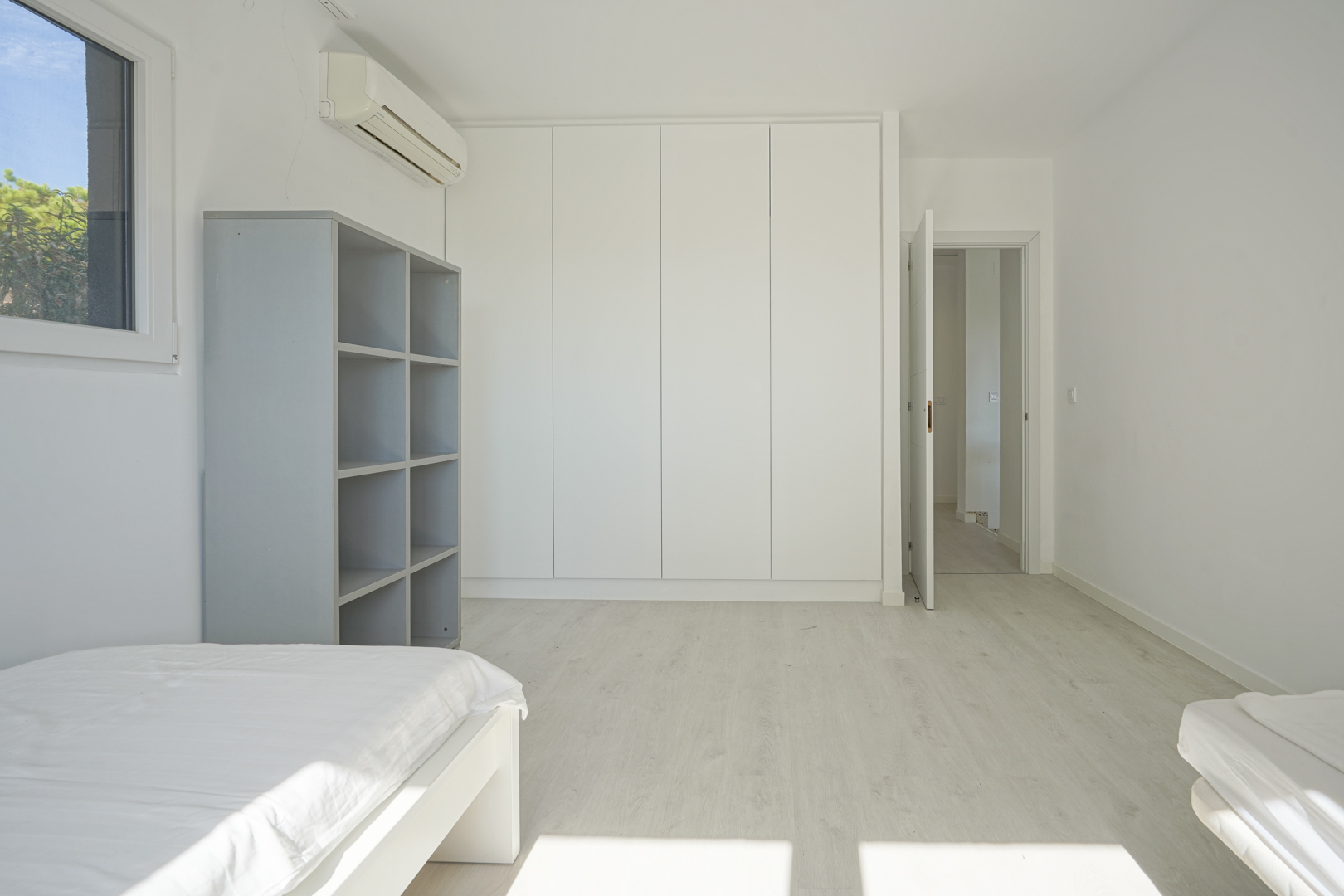 Chambre lumineuse avec placards intégrés blancs, sol en stratifié clair, climatisation murale et étagère grise, offrant un design épuré et fonctionnel.
