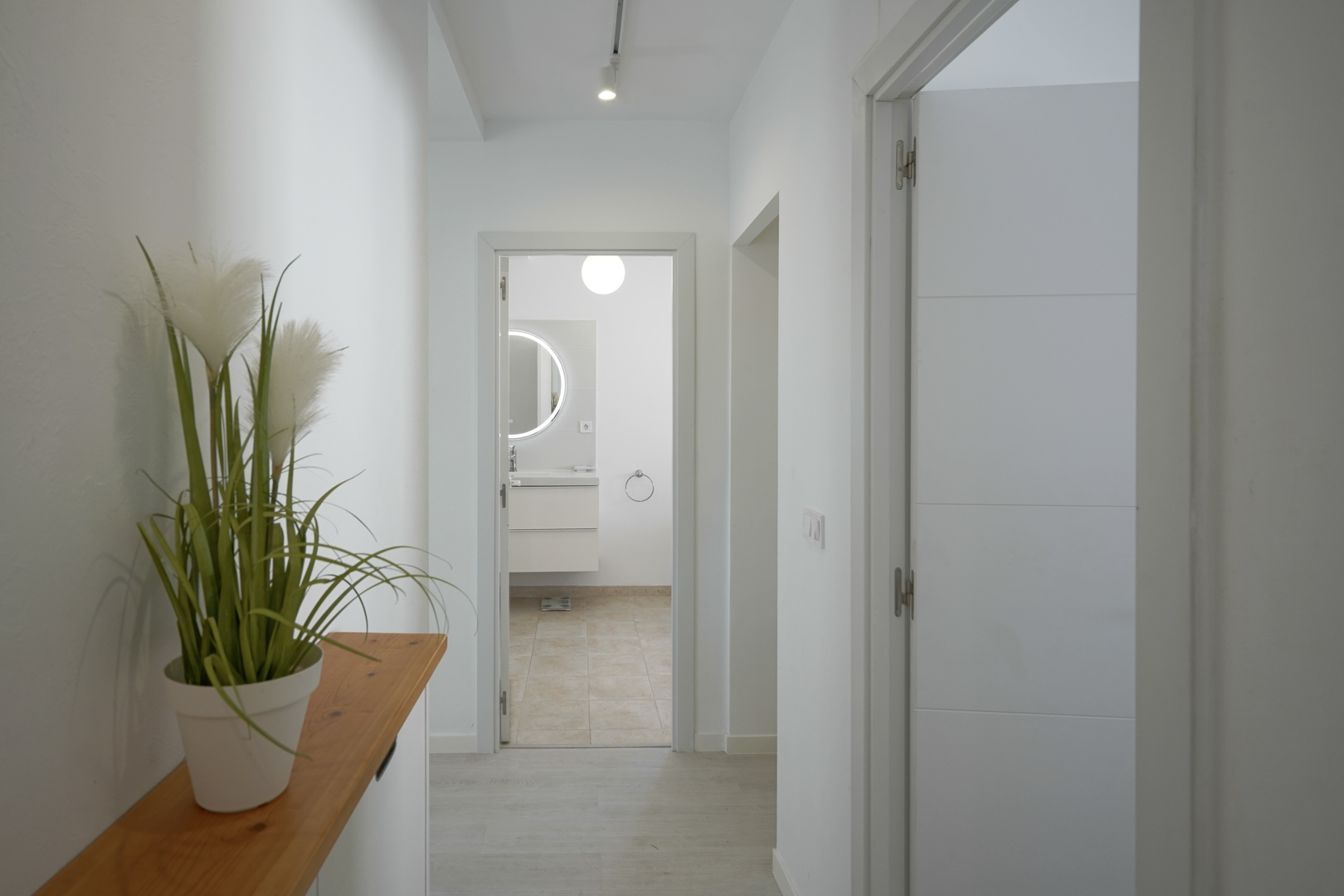 Couloir lumineux aux murs blancs et sol clair menant à une salle de bain moderne avec miroir circulaire rétroéclairé et meuble suspendu.