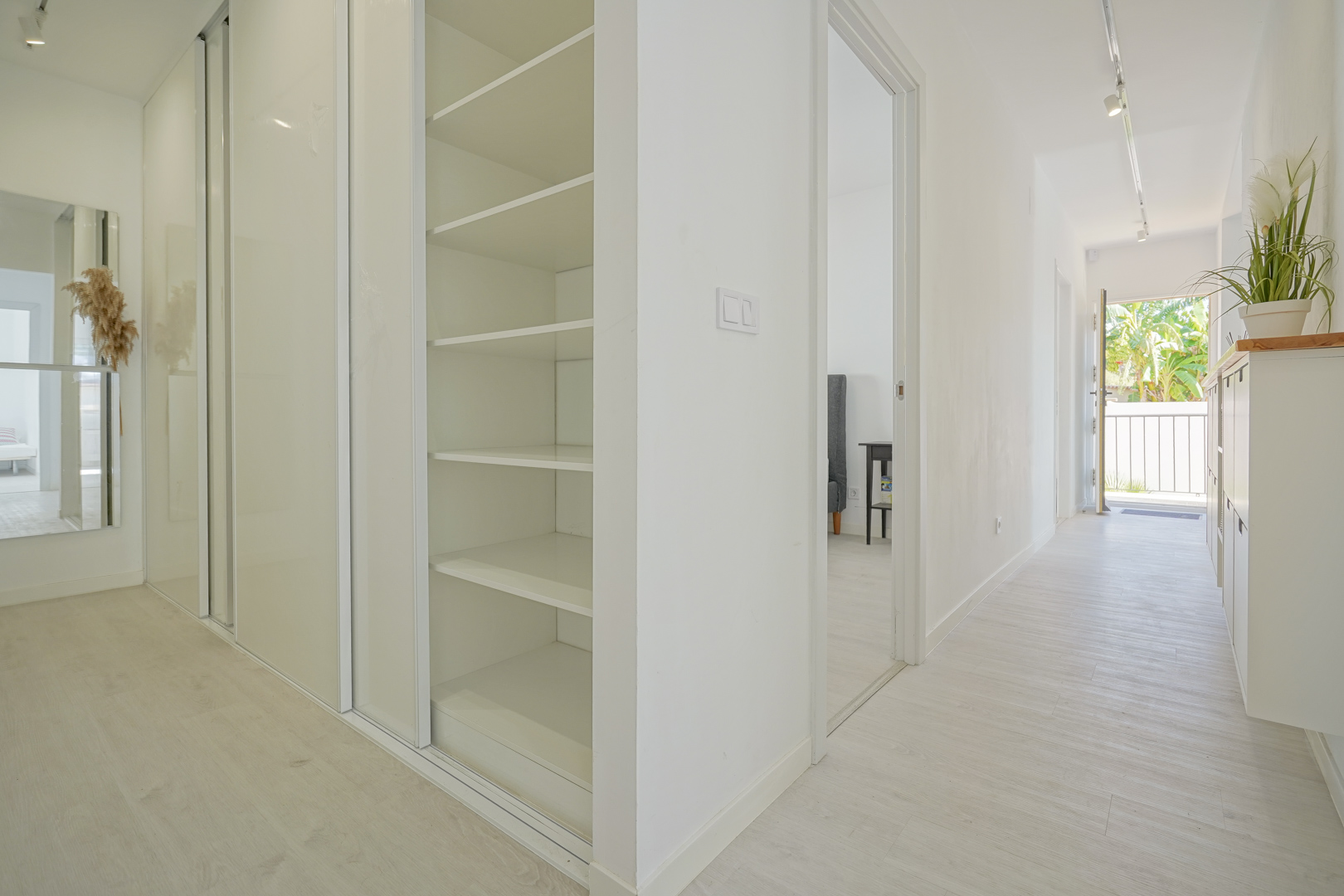 Couloir lumineux avec placards intégrés blancs, étagères ouvertes et sol en parquet clair menant vers l'entrée de la villa.
