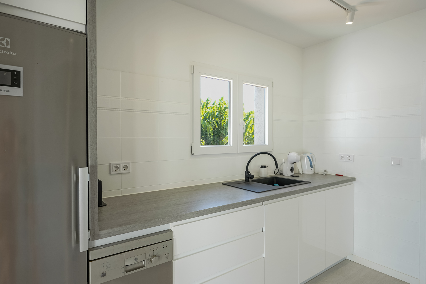 Cuisine fonctionnelle avec placards blancs sans poignées, plan de travail gris, évier noir et appareils intégrés sous une fenêtre lumineuse.