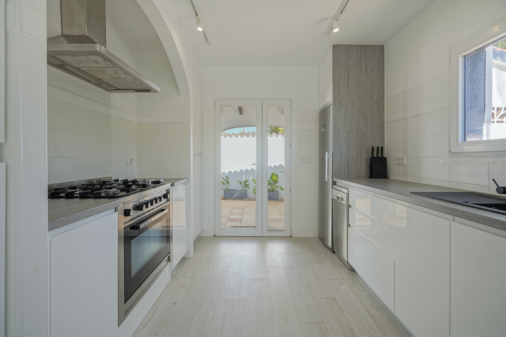 Cuisine moderne avec placards blancs brillants, plans de travail gris et électroménager en inox. Elle dispose d'une gazinière et d'un accès direct à la terrasse.