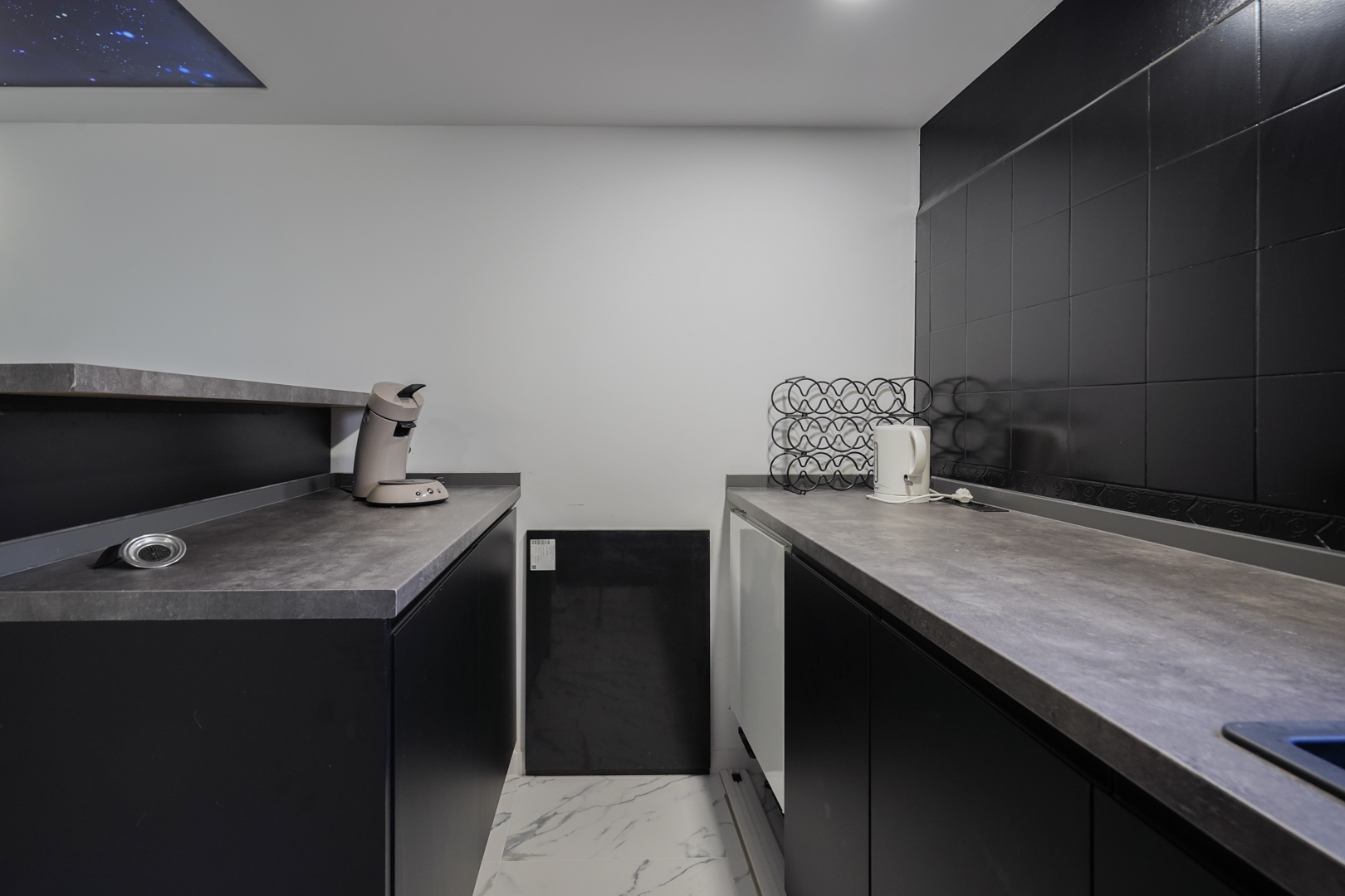 Espace cuisine secondaire avec placards noirs, plans de travail gris et sol effet marbre. Un panneau de plafond décoratif ciel étoilé complète l'aménagement.