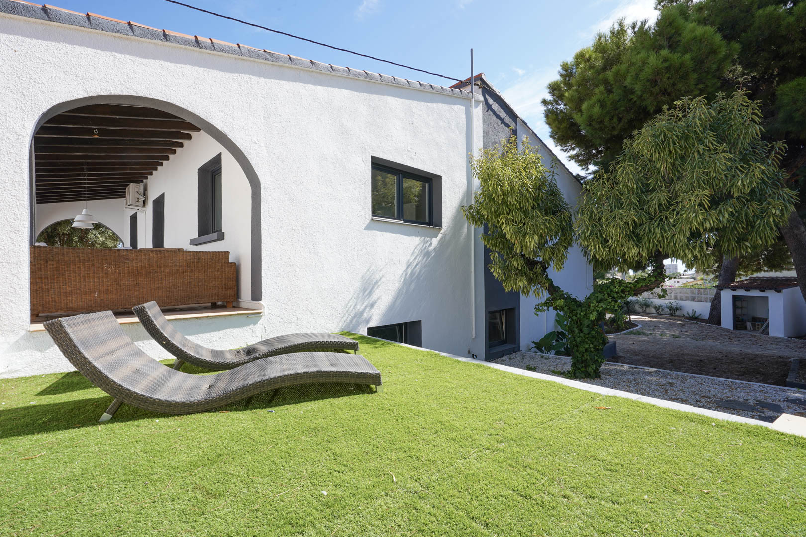 Espace de pelouse surélevé avec chaises longues en osier devant la façade blanche et une terrasse couverte aux poutres en bois sombre.