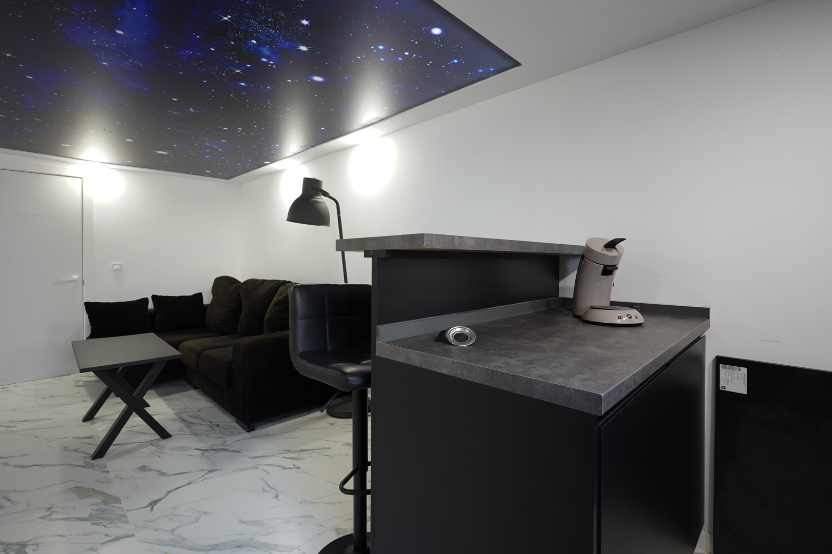 Espace détente avec bar à café, canapé sombre et sol effet marbre, doté d'un plafond rétroéclairé unique représentant un ciel étoilé.