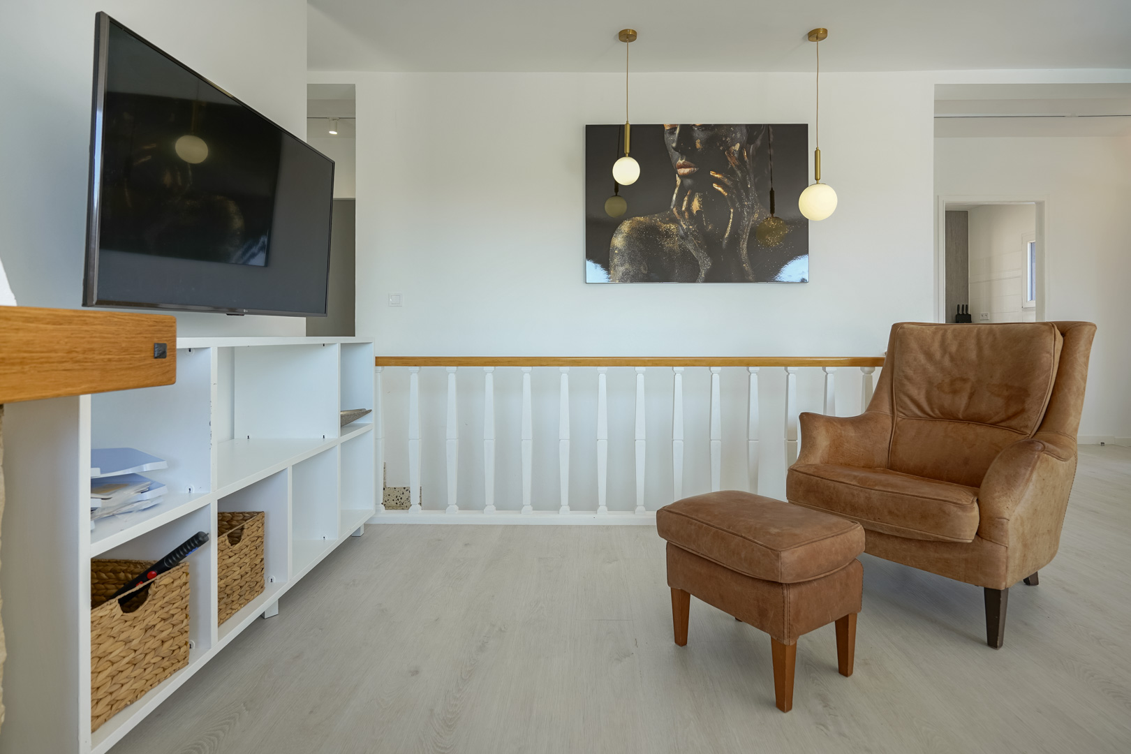 Espace détente avec fauteuil en cuir et repose-pieds, étagère blanche et balustrade en bois. Éclairage suspendu moderne et sol stratifié aux tons clairs.