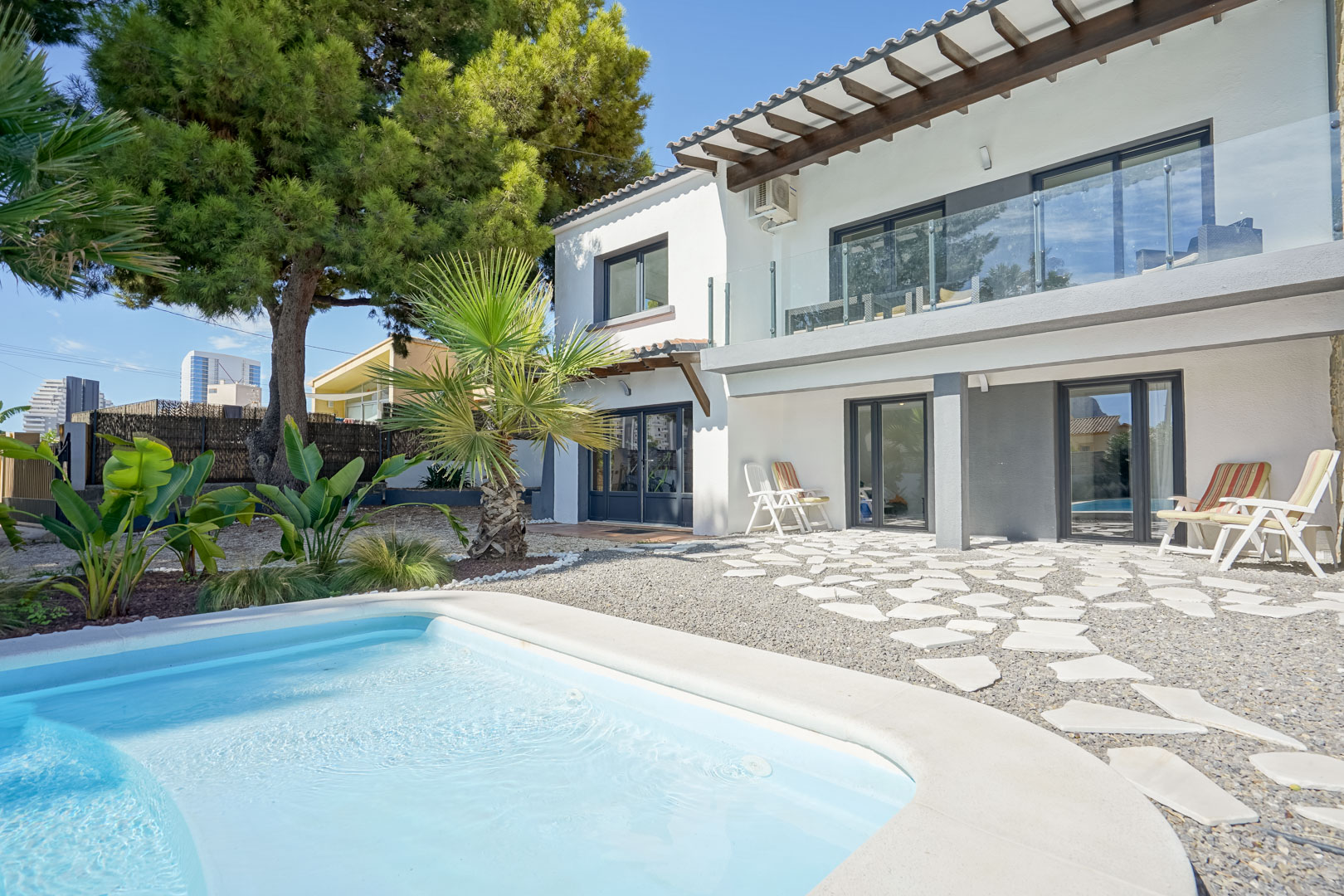 Façade arrière de la villa avec piscine, terrasse supérieure à garde-corps en verre et poutres en bois, et patio en gravier avec dalles irrégulières.