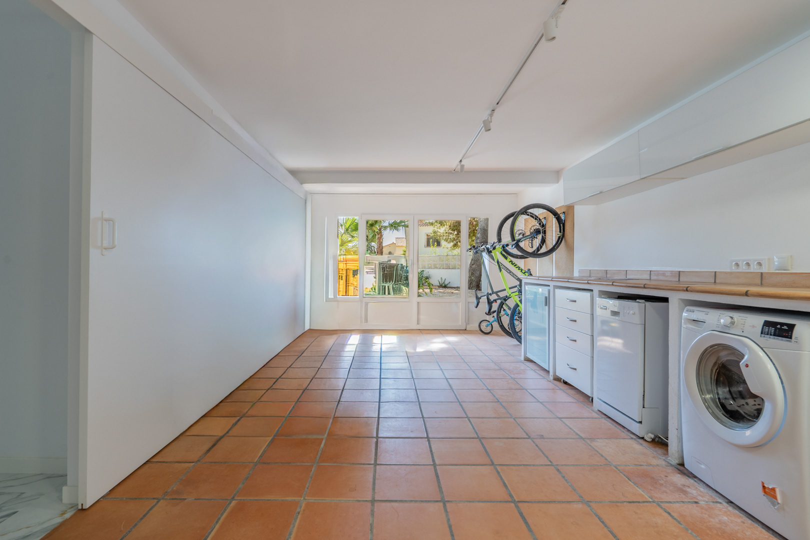 Garage aménagé en buanderie et espace de rangement avec sol en terre cuite, plan de travail carrelé, électroménager et accès vitré vers l'extérieur.