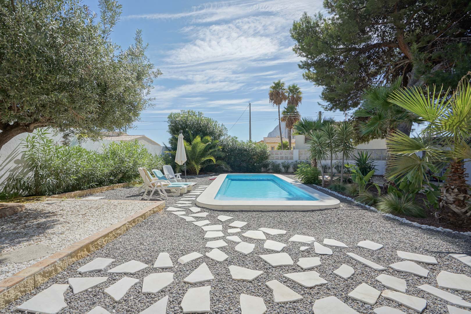 Piscine rectangulaire entourée d'un jardin méditerranéen avec oliviers et palmiers. Comprend un chemin en pierres irrégulières et une zone de bronzage.