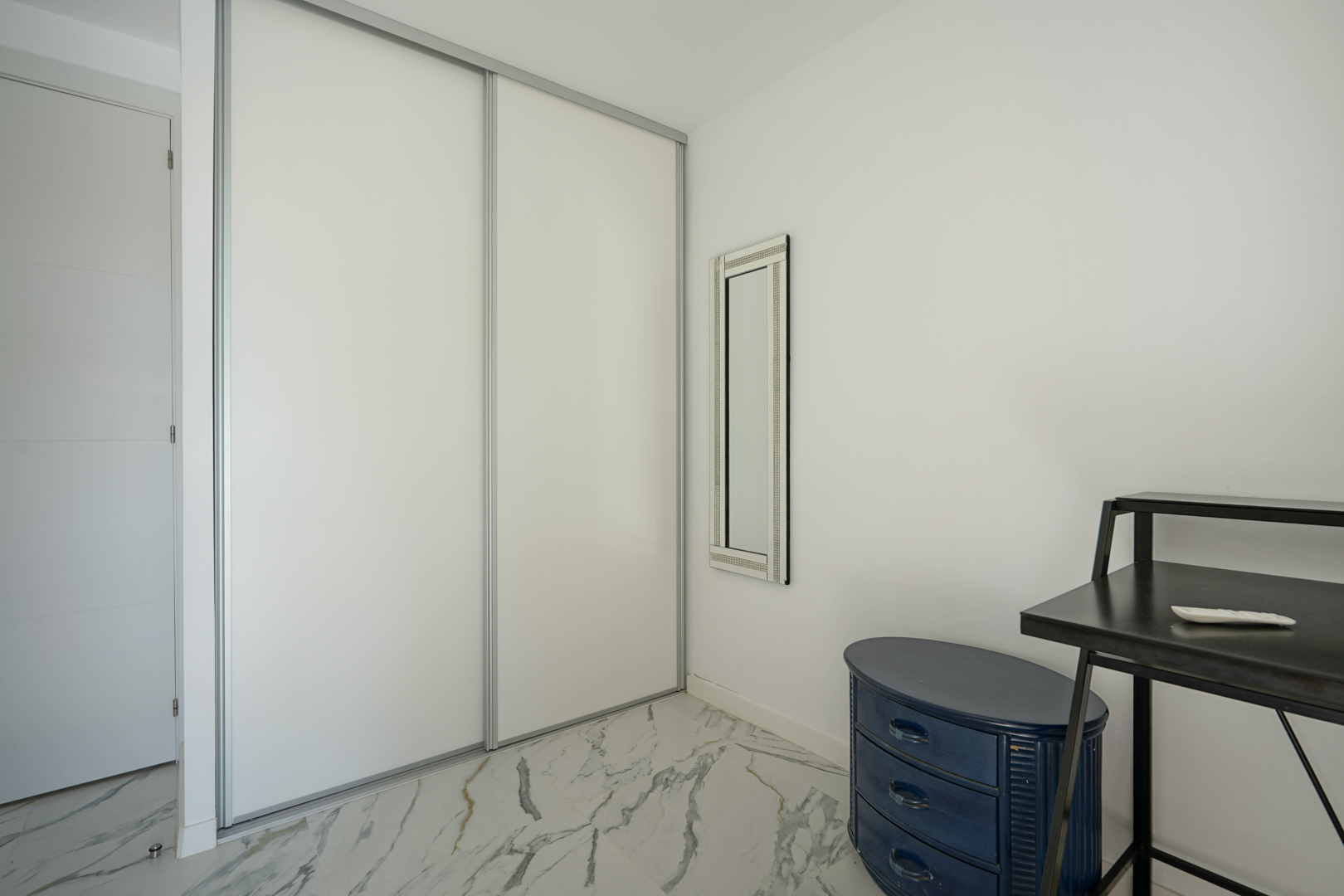 Placard intégré avec portes coulissantes blanches, sol en marbre veiné, miroir vertical et commode ovale bleue dans un espace épuré.