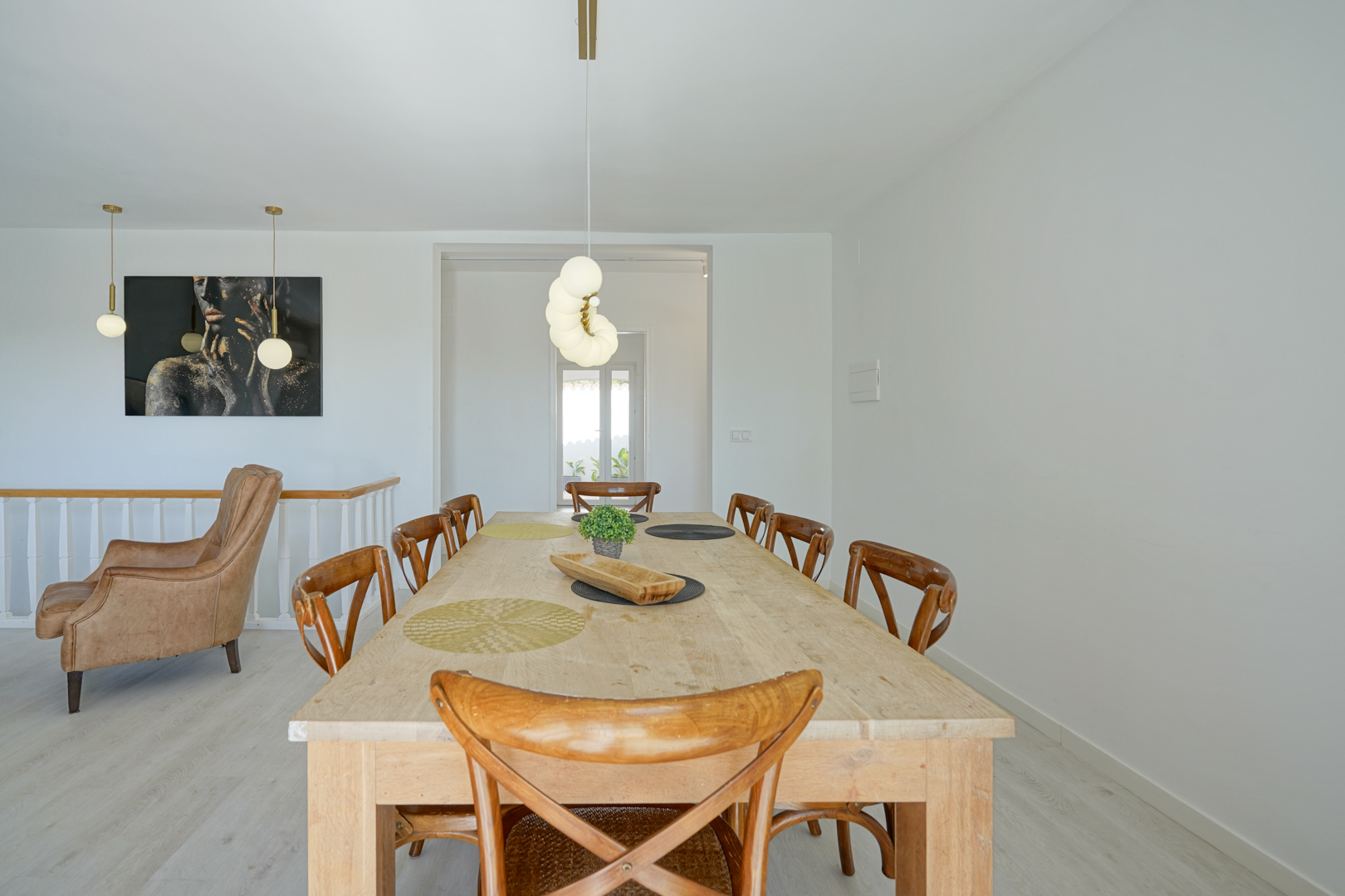 Salle à manger avec une grande table en bois clair, des chaises à dossier croisé et des suspensions sphériques modernes. Un fauteuil en cuir et une peinture complètent l'espace.