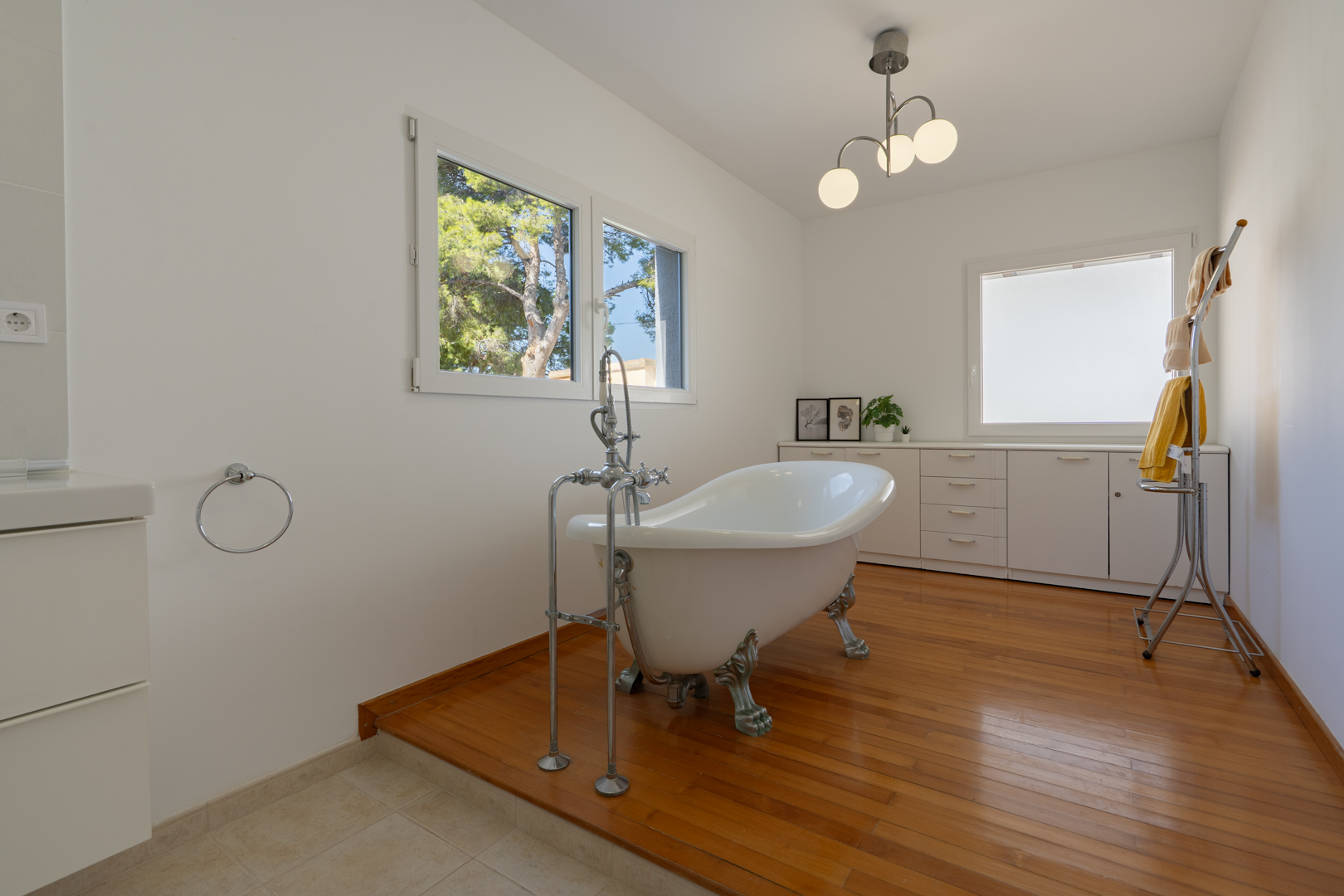 Salle de bains avec baignoire îlot à pattes de lion sur parquet, meubles de rangement blancs et fenêtres offrant une vue sur la végétation environnante.