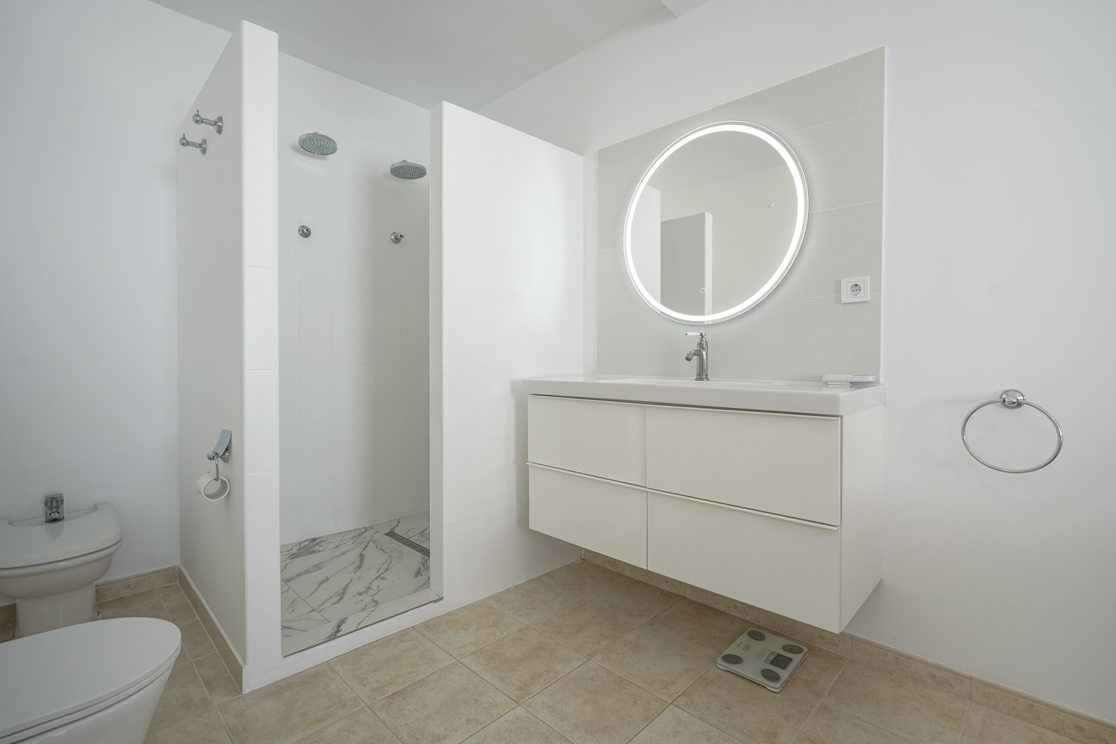 Salle de bains moderne avec meuble vasque suspendu blanc, miroir circulaire rétroéclairé et une grande douche à l'italienne double au sol effet marbre.